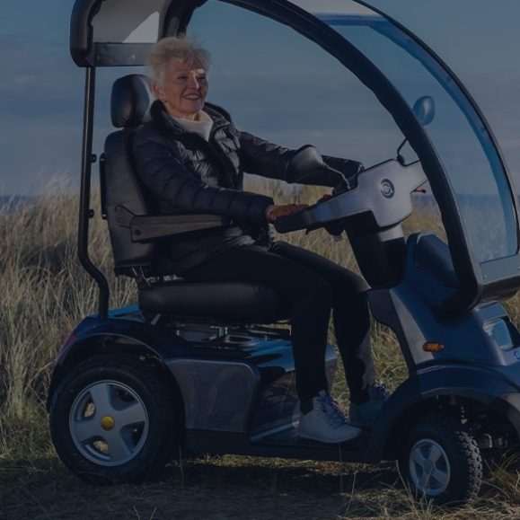 An elderly person sitting on a mobility scooter on a grassy area.